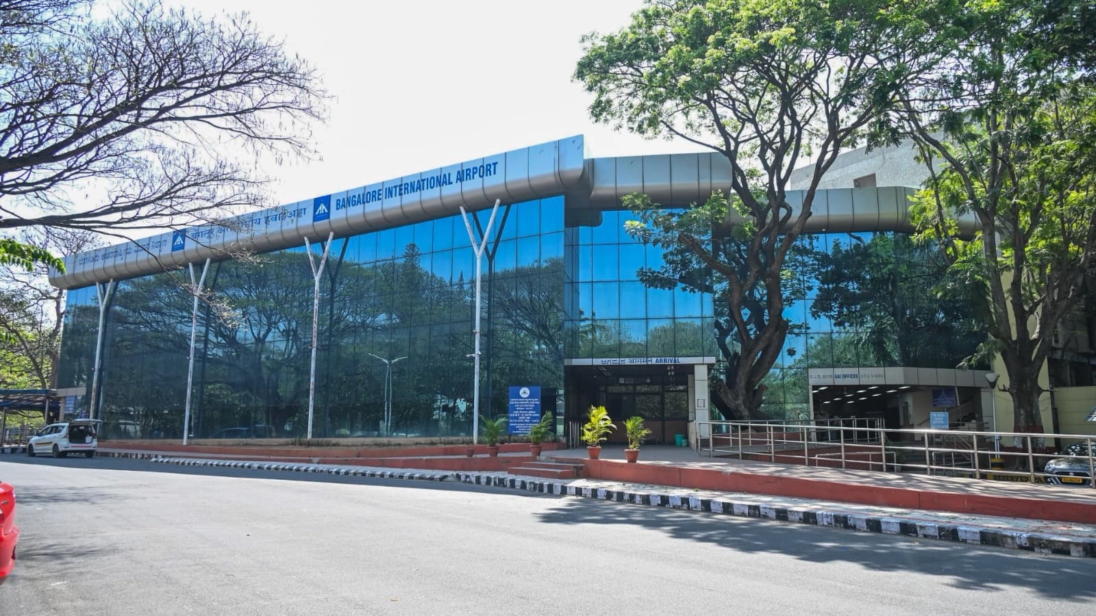 The glass-fronted arrival and departure lounge at Bangalore airport, its silver fascia carrying the airport's name, rain trees reflected in the glazing