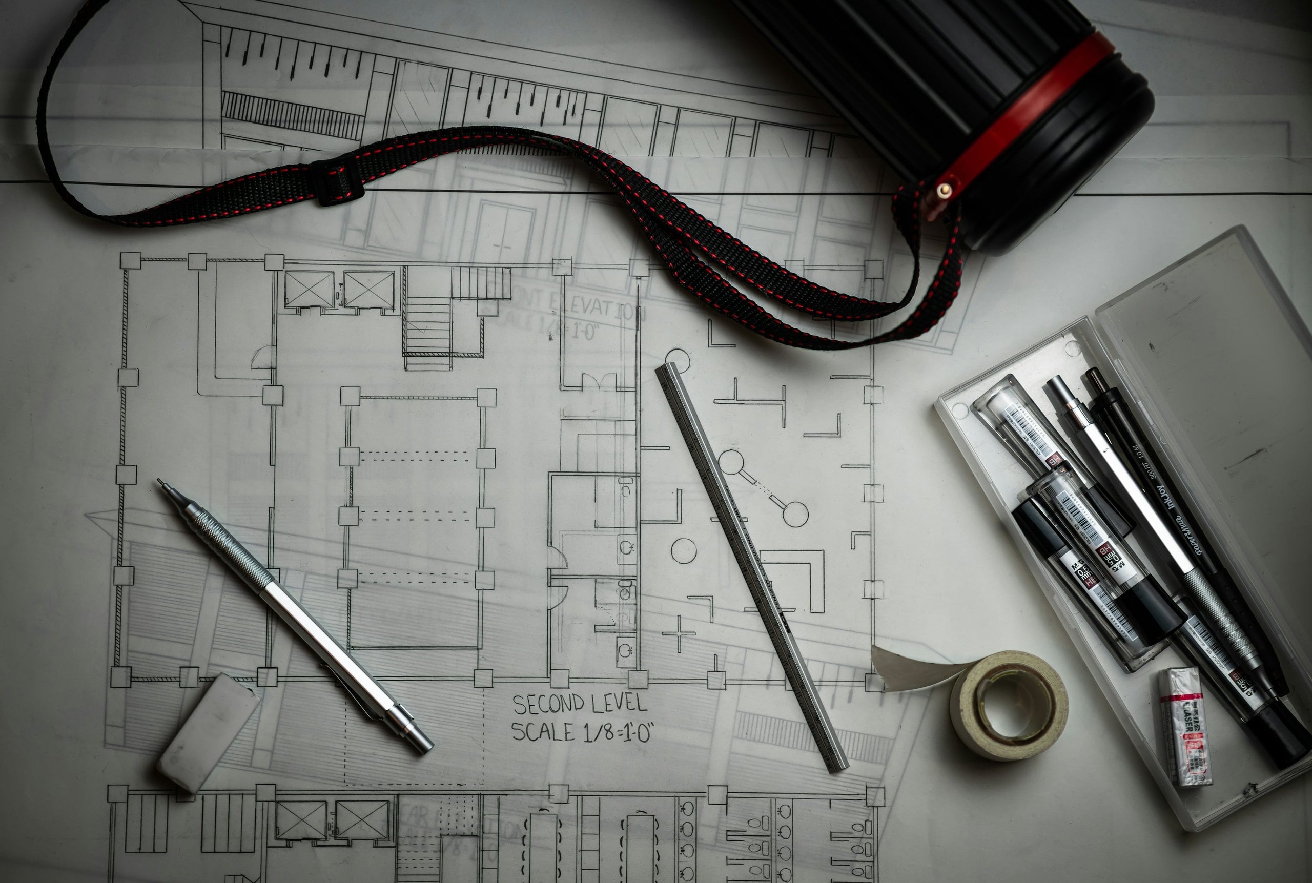 A floor plan on a drawing board, with a scale rule, drafting pens and a drawing tube laid across it