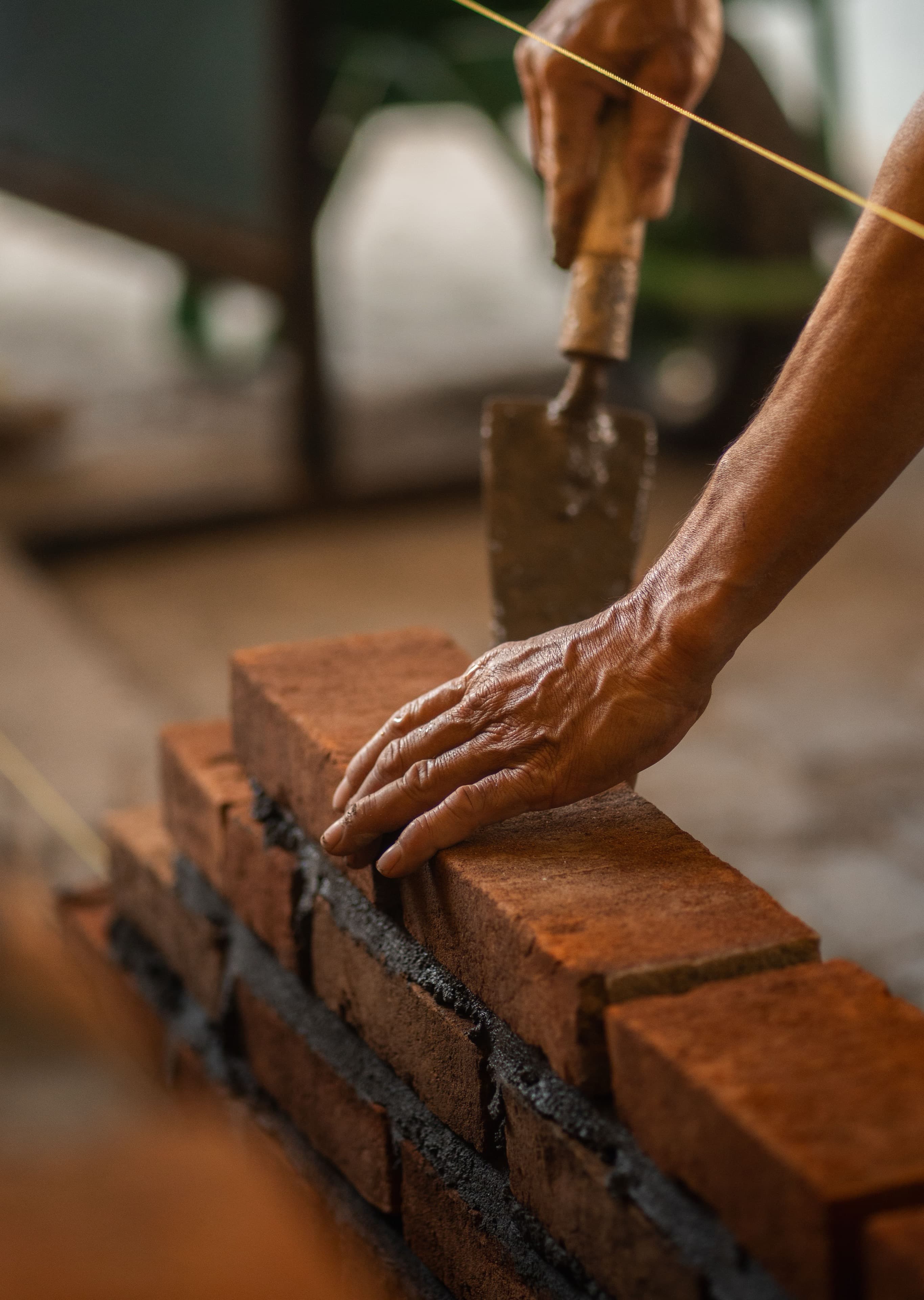A mason's hands bedding a brick down onto fresh mortar against the line, trowel still in the other hand