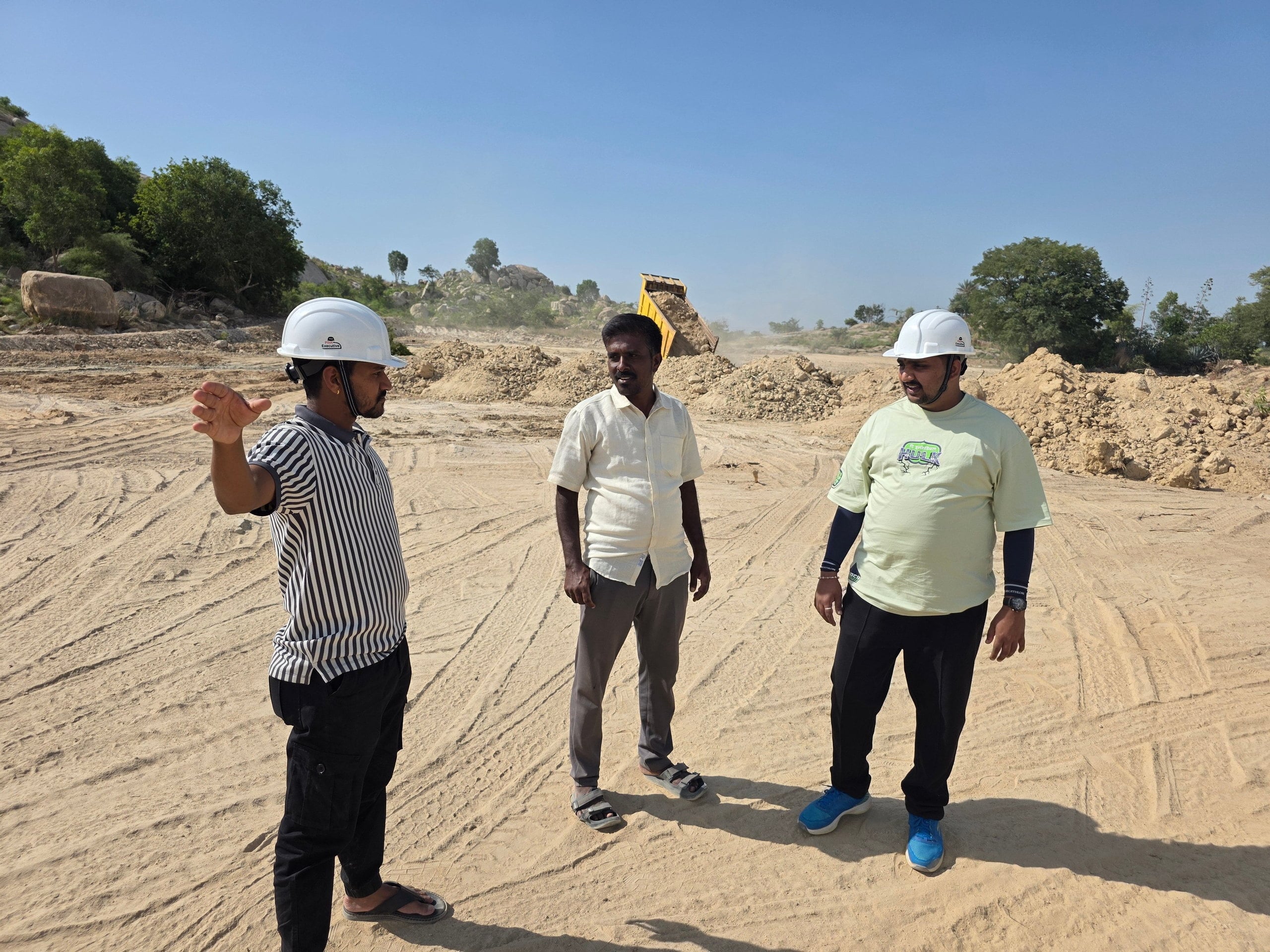 Three men standing together on a cleared plot, two of them in hard hats, an excavator tipping earth behind them