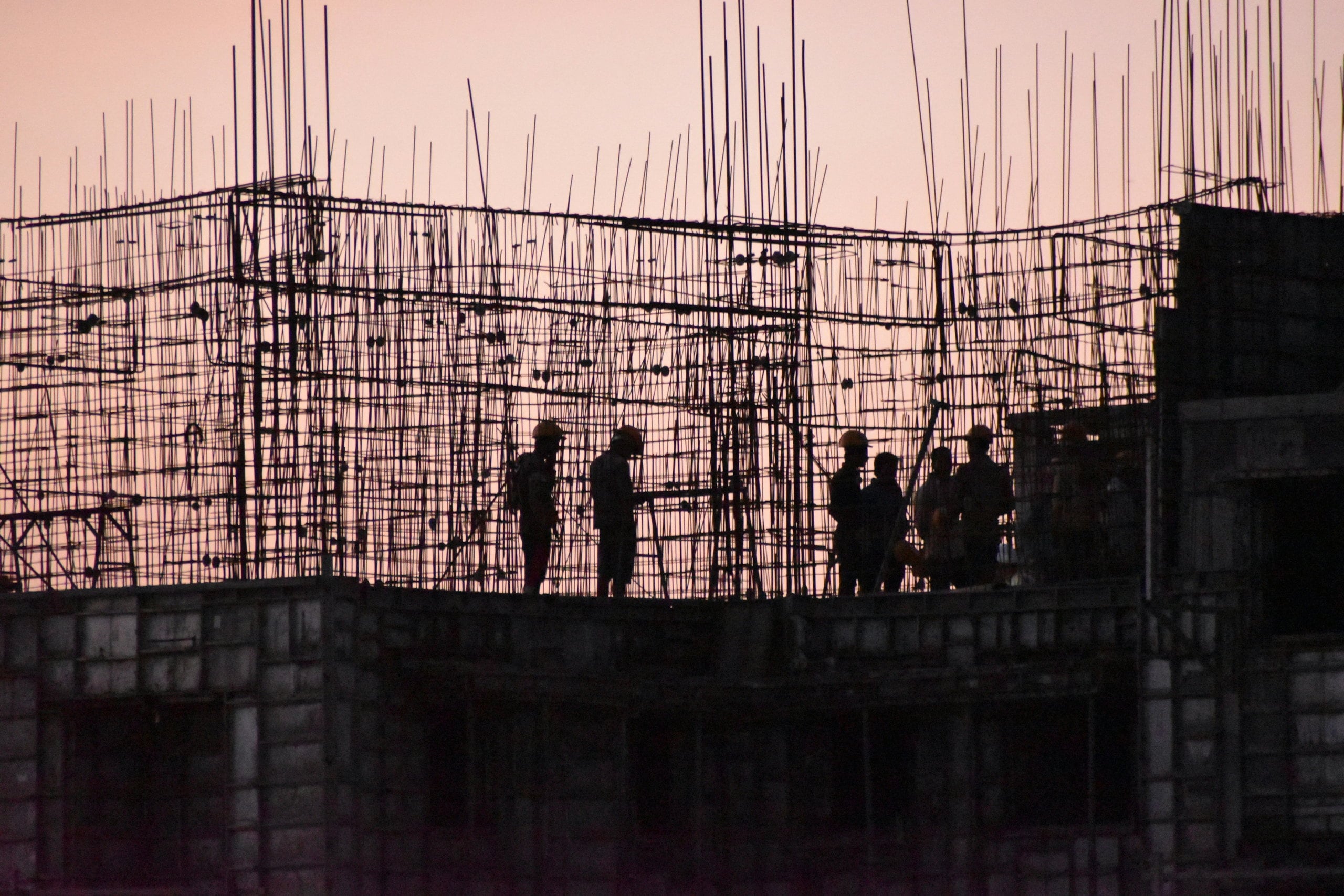 Workers silhouetted at dusk against a pink sky, on the reinforcement cage of a floor going up