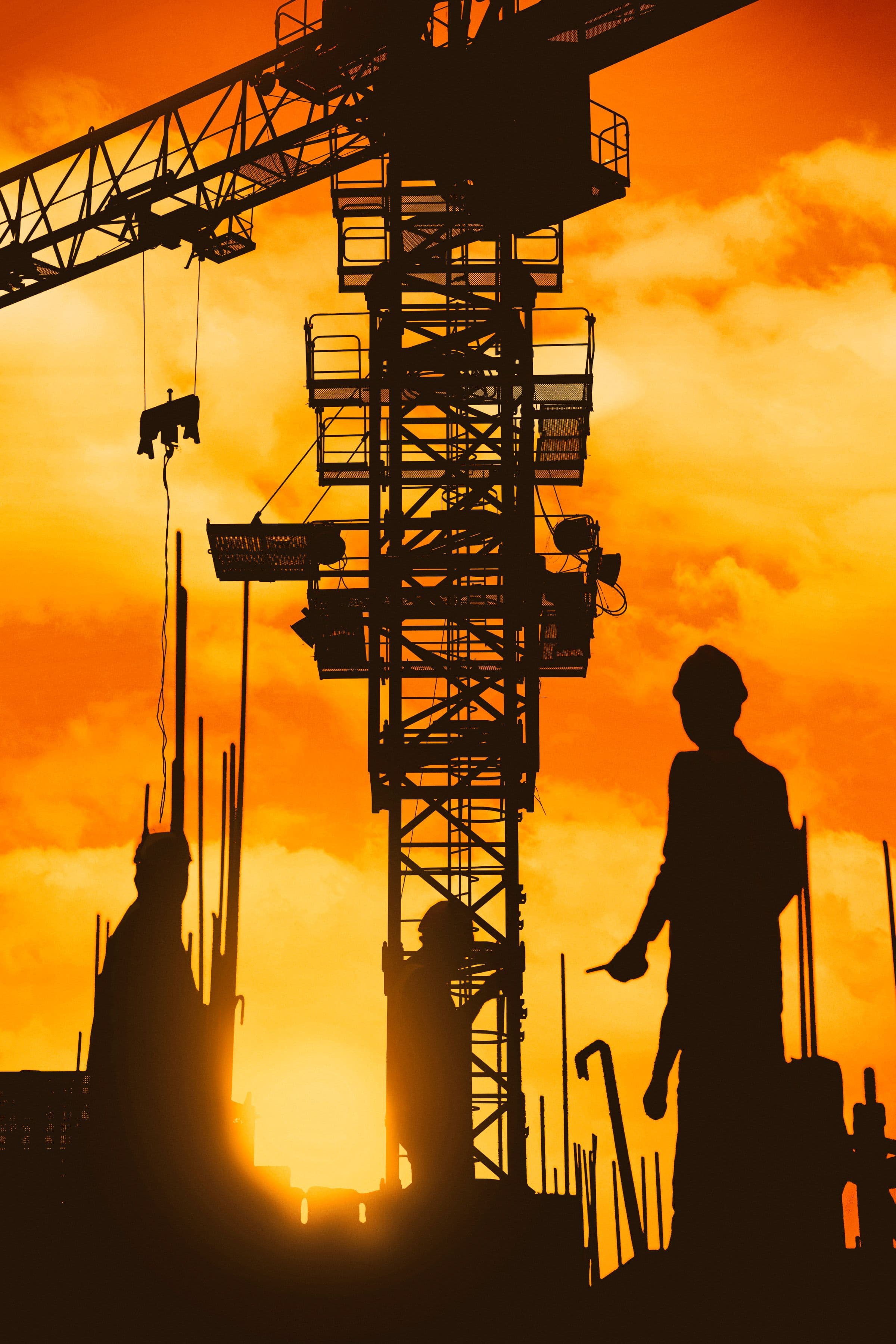 A tower crane and two workers in silhouette against a burning orange sky, reinforcement bars rising around them