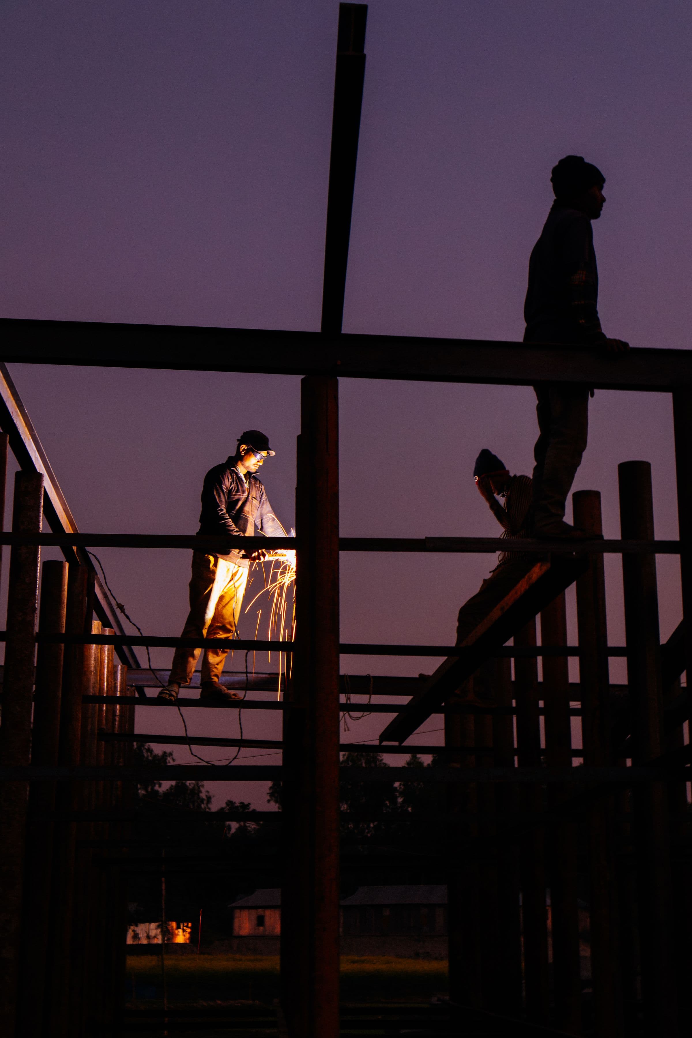 A welder at work on a steel frame at dusk, sparks falling from the torch, a second worker on the beam above