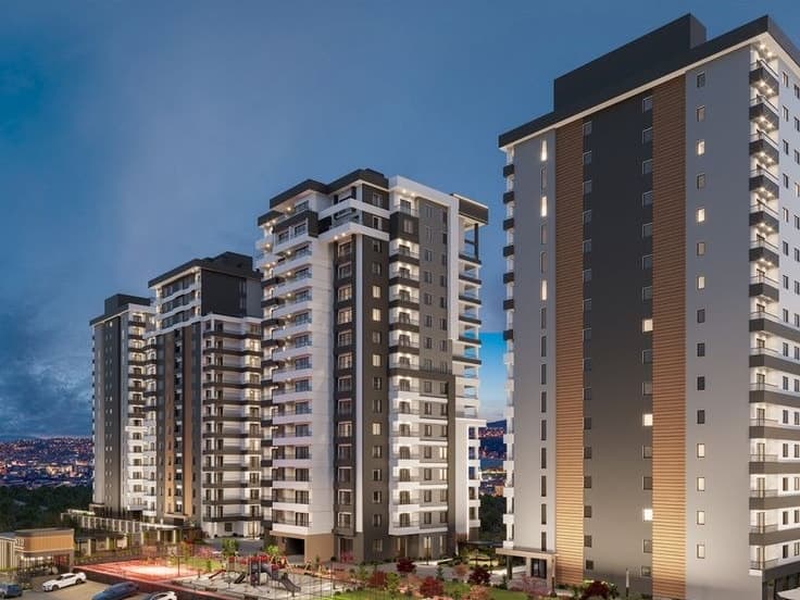 Four residential towers at dusk in white, charcoal and timber cladding, lit windows over a landscaped court with a play area and cars parked along the road